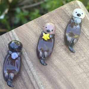 May include: Three small, glossy, sculpted otter figurines in dark brown. The left otter holds a light purple shell, the center otter holds a bright yellow star, and the right otter has a lighter tan head and holds a small gray circular object.