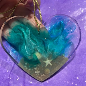 May include: A heart-shaped resin ornament with a blue and green swirl design, reminiscent of ocean waves. The bottom of the ornament features a sandy beach with a starfish and seashell.