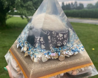 May include: A clear glass pyramid filled with layers of sand, seashells, and a small dog collar. The top layer contains white fur.