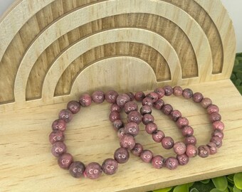 May include: Three pink gemstone bracelets with a repeating pattern of round beads. The bracelets are stacked on top of each other on a wooden rainbow-shaped stand.