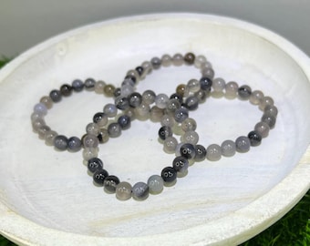 May include: Three gray and black agate beaded bracelets stacked on a white plate. The beads are round and have a natural, polished finish.