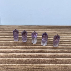 May include: Five amethyst crystal points, each with a pointed tip and a clear, translucent body. The crystals are arranged in a row on a wooden surface.