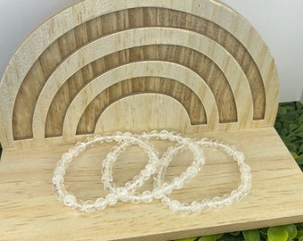 May include: Three clear quartz crystal beaded bracelets stacked on a wooden rainbow-shaped display.