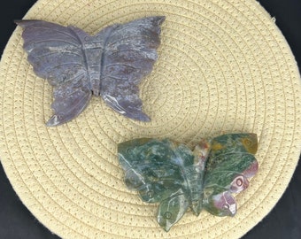 May include: Two stone butterfly sculptures on a beige woven surface. The butterfly on the left is a dark gray color, while the butterfly on the right is a multi-colored green, brown, and red.