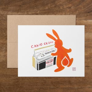 May include: Greeting card featuring an orange rabbit playing a Casio CK200 keyboard. The keyboard is depicted in gray, black, and gold, with the text "CASIO CK200" in red. The card is on a white background, with a brown envelope.