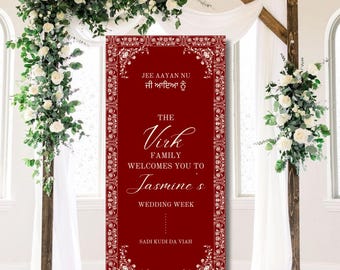May include: A red wedding welcome sign with white ornate borders and text. The text reads "Jee Aayan Nu Ji Aaiya Nu" in Punjabi, "The Virk Family Welcomes You To Jasmine's Wedding Week" in English, and "Sadi Kudi Da Viah" in Punjabi.