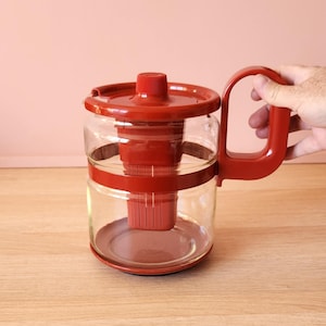 May include: A clear glass coffee maker with a red plastic lid, handle, and internal filter. The coffee maker has a cylindrical shape with a red band around the middle. The handle is a curved, open design. The coffee maker is designed for brewing coffee.