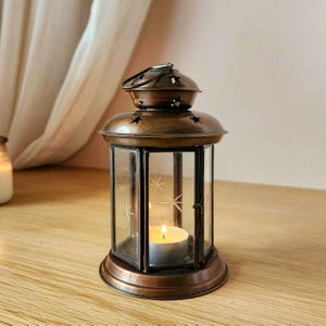 May include: A hexagonal metal lantern with a dark bronze finish and a hinged door. The top features cut-out star shapes, while the glass panels are etched with star patterns. A small, lit white tea light candle sits inside the lantern base.
