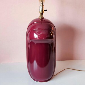 May include: A burgundy-colored ceramic table lamp with a glossy finish. The lamp has a rounded, elongated shape with vertical grooves and a brass-colored light fixture. A light bulb is visible at the top, and a brown electrical cord extends from the base.
