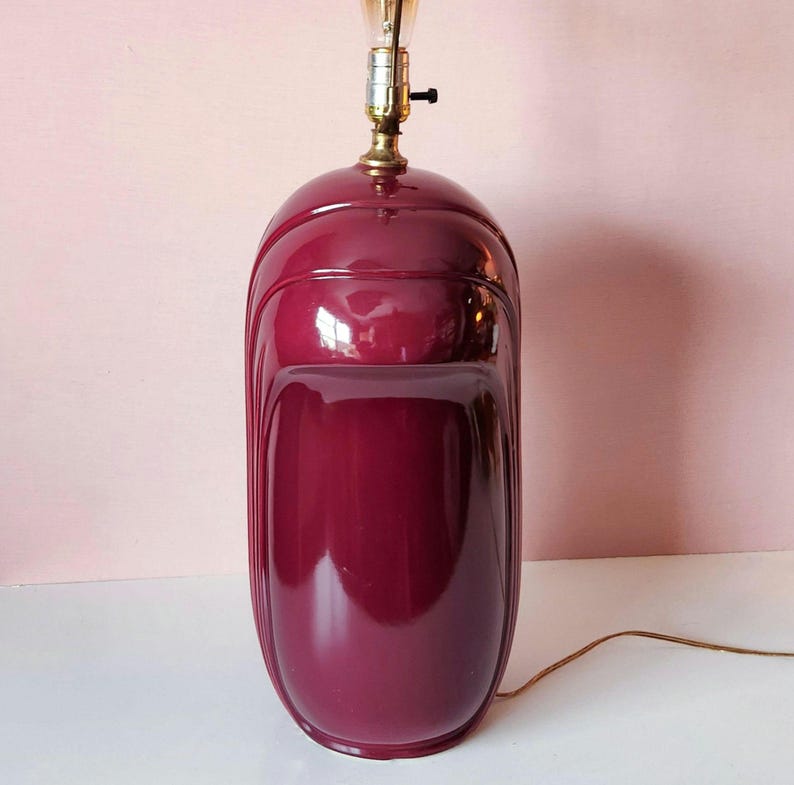 May include: A burgundy-colored ceramic table lamp with a glossy finish. The lamp has a rounded, elongated shape with vertical grooves and a brass-colored light fixture. A light bulb is visible at the top, and a brown electrical cord extends from the base.