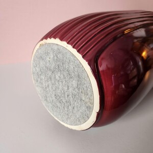 May include: A maroon-colored ceramic vase with a textured, ribbed design. The vase has a white rim and a gray felt base. The glossy finish reflects light, highlighting the curves and details of the vessel.
