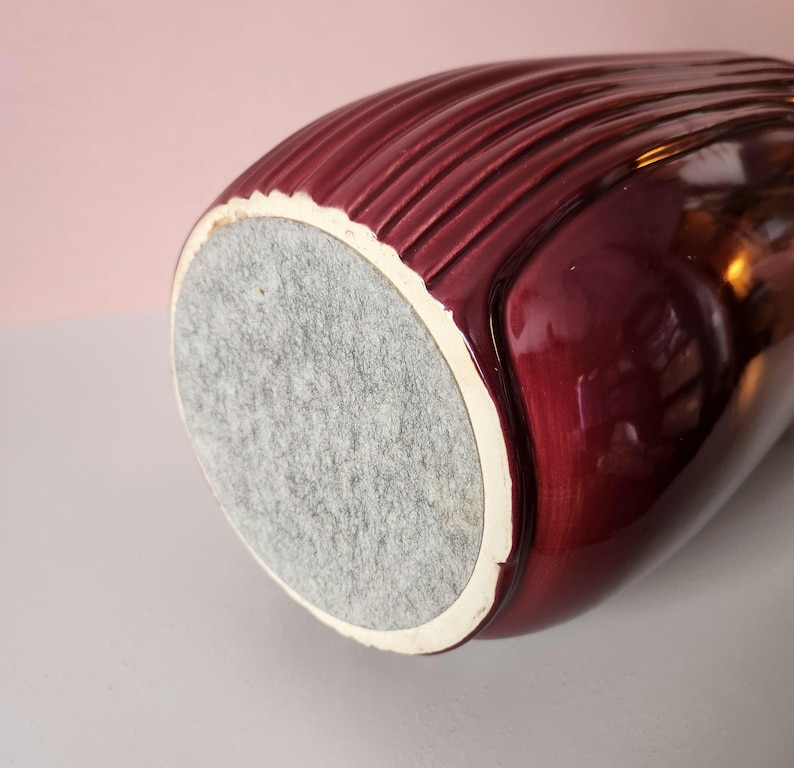 May include: A maroon-colored ceramic vase with a textured, ribbed design. The vase has a white rim and a gray felt base. The glossy finish reflects light, highlighting the curves and details of the vessel.