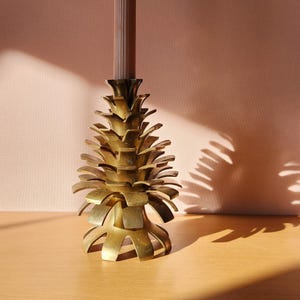 May include: A gold metal candle holder shaped like a pine cone. The candle holder is on a wooden surface.