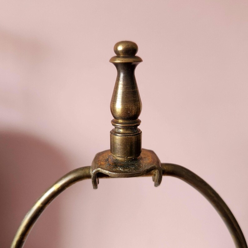 May include: Close-up of a vintage brass lamp finial with a decorative, turned design. The finial is mounted on a curved brass frame, with a warm, aged patina. The background is a soft, muted pink.