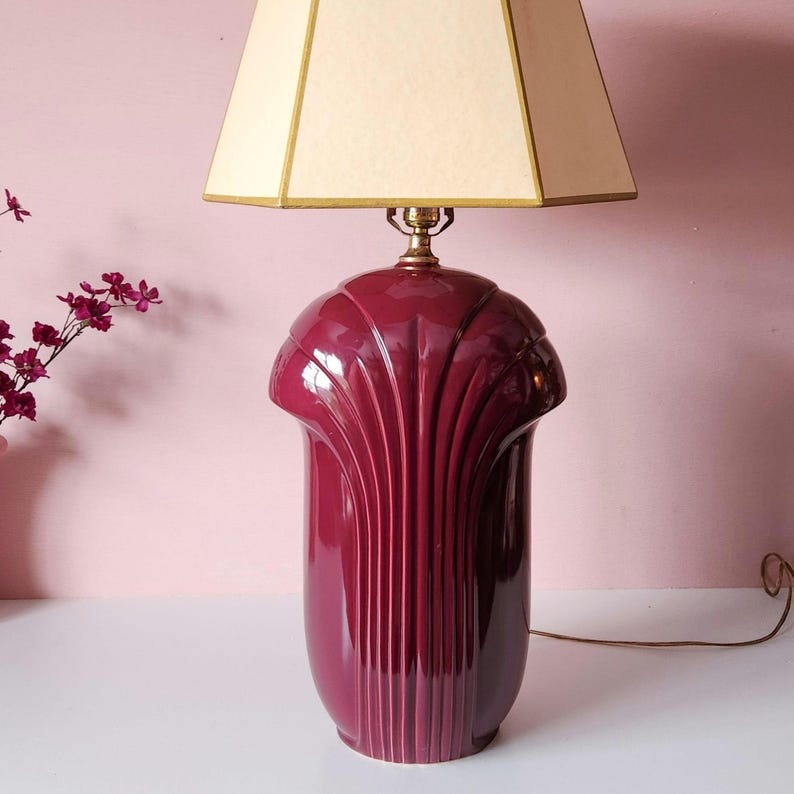May include: A burgundy ceramic table lamp with a cream-colored lampshade. The lamp base has a fluted design and a brass-colored neck. The lampshade is a square shape with gold trim. The lamp is on a white surface against a pink background.