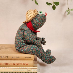 May include: A green frog plush toy wearing a straw hat and a red bandana. The frog is sitting on a stack of books. The books have the titles "Street", "Harper", "Of the Old Post Box", and "Tree".