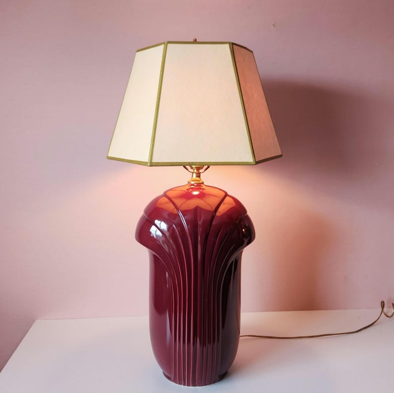 May include: A burgundy table lamp with a cream-colored, geometric lampshade. The lamp base has a fluted design and a glossy finish. The lampshade is trimmed with a gold border. The lamp is plugged into an electrical outlet.
