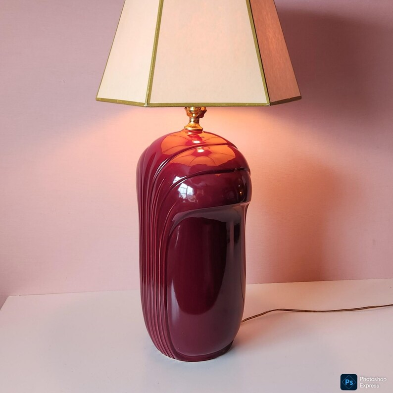 May include: A burgundy ceramic table lamp with a cream-colored lampshade. The lamp base has a glossy finish with vertical grooves on the sides. The lampshade is square and trimmed with gold. The lamp is on a white surface.