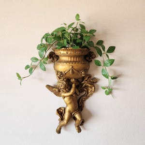 May include: A gold-colored wall planter featuring a cherub holding a decorative urn filled with green leafy plants. The urn has ornate detailing and a classic design. The cherub has wings and is attached to a wall mount.