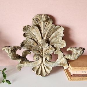 May include: A decorative wall sconce with a distressed white and gold finish. The sconce features a floral design with intricate details and two candle holders.