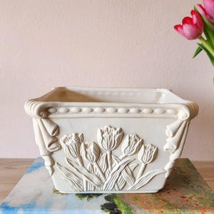 May include: Cream-colored rectangular planter with embossed tulip design. The planter has a decorative border and stylized corner accents. The planter is sitting on a colorful surface, with pink tulips in the background.