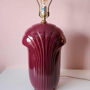 May include: A vintage burgundy table lamp with a brass neck and a decorative, fan-like design. The lamp has a bulb installed and a brown electrical cord. The lamp is on a white surface against a pink background.