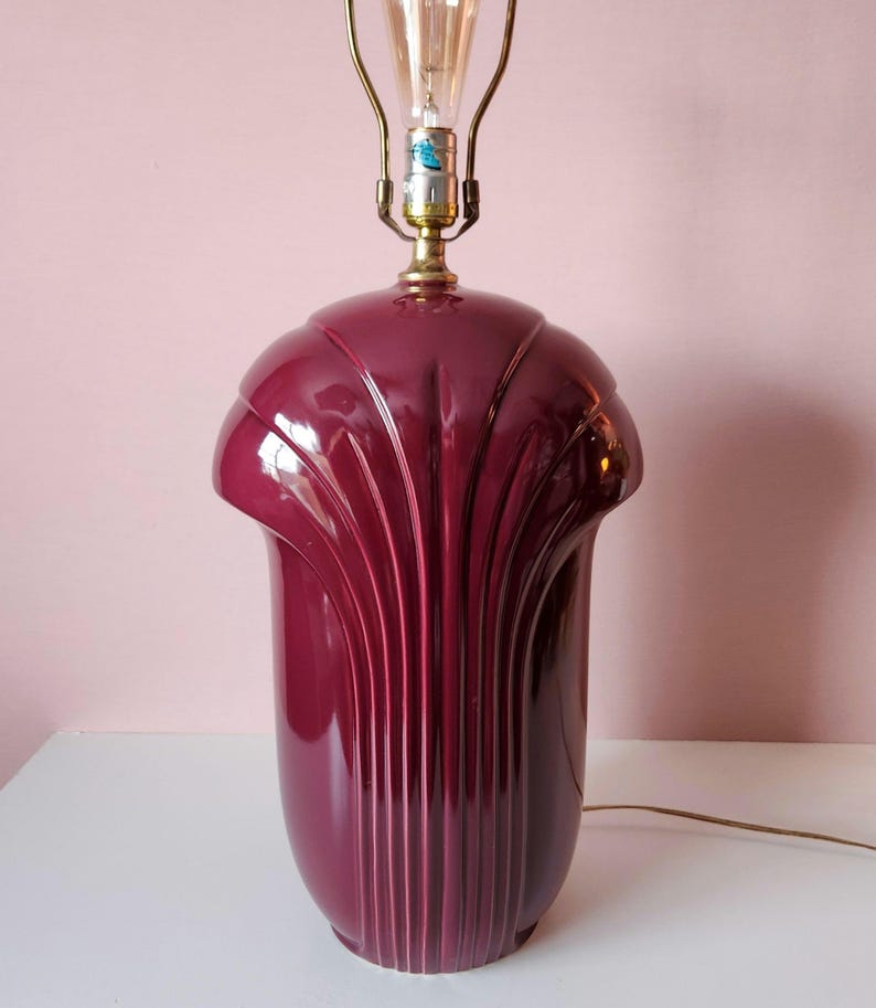 May include: A vintage burgundy table lamp with a brass neck and a decorative, fan-like design. The lamp has a bulb installed and a brown electrical cord. The lamp is on a white surface against a pink background.