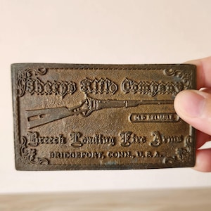 May include: A rectangular, bronze-colored plaque with embossed text and a rifle illustration. The text reads "Sharps Rifle Company," "Breech Loading Fire Arms," and "Old Reliable." The plaque is from Bridgeport, Conn., U.S.A.