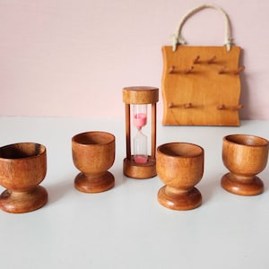 May include: Four wooden egg cups and a wooden hourglass with pink sand. The egg cups are brown and have a rounded base. The hourglass is on a white surface.