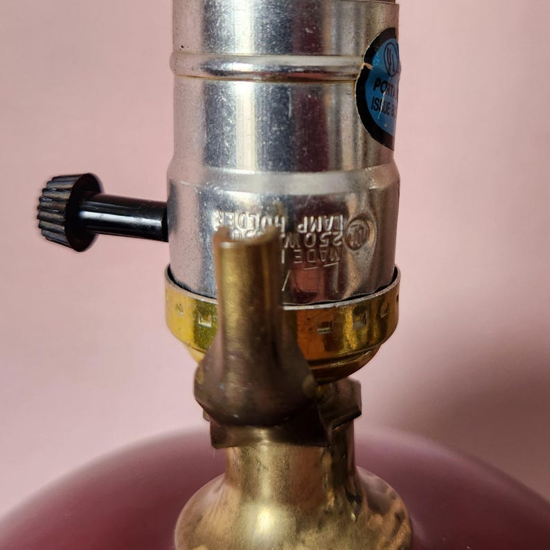 May include: Close-up of a lamp base with a silver lamp holder and a black knob. The base is a deep red color, and the lamp holder has a gold-colored ring. A blue and white sticker is visible on the lamp holder.
