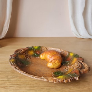 May include: An oval-shaped decorative tray with a wood grain texture, featuring sculpted grape clusters and leaves in shades of green, yellow, and brown around the rim. A single golden-brown croissant-shaped object rests in the center.
