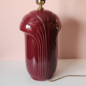 May include: A burgundy-colored Art Deco style table lamp. The lamp has a rounded top and a cylindrical base with vertical, curved grooves. The lamp has a brass-colored socket and a brown electrical cord.