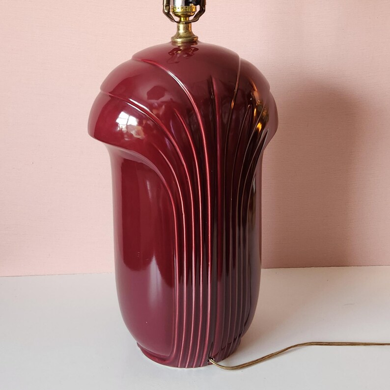 May include: A burgundy-colored Art Deco style table lamp. The lamp has a rounded top and a cylindrical base with vertical, curved grooves. The lamp has a brass-colored socket and a brown electrical cord.