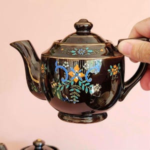 May include: A black ceramic teapot with a lid, spout, and handle. The teapot is decorated with colorful floral patterns in blue, orange, and white, accented with gold trim. The teapot is held by a hand.