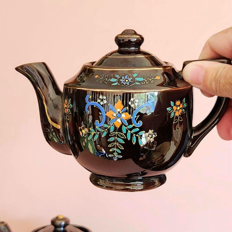 May include: A black ceramic teapot with a lid, spout, and handle. The teapot is decorated with colorful floral patterns in blue, orange, and white, accented with gold trim. The teapot is held by a hand.