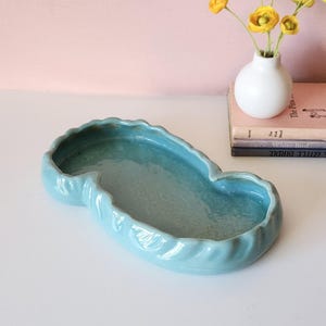 May include: A light blue ceramic decorative dish with a wavy, scalloped edge. The dish has a unique, elongated shape and a glossy finish. The dish is empty and sits on a white surface. A small white vase with yellow flowers and a stack of books are in the background.