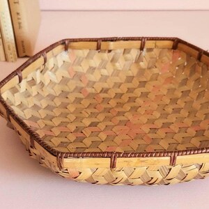 May include: A hexagonal woven basket featuring a light tan and brown geometric pattern. The basket has a reinforced dark brown rim and a flat base, constructed from thin strips of natural material with a visible interlocking weave design.