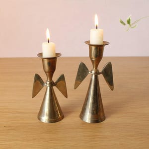 May include: Two brass candle holders shaped like angels with conical bodies and outstretched wings. Each holder features a cylindrical top section holding a lit white pillar candle, creating a warm glow from the metallic surfaces.
