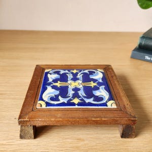 May include: A square wooden trivet featuring a central ceramic tile with a blue, white, and yellow floral-inspired pattern. The frame is constructed from medium-toned wood with four small feet at the corners, providing a raised base for the tile.
