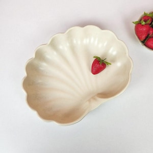 May include: A cream-colored, shell-shaped serving dish with a single red strawberry. The dish has a ribbed design and a scalloped edge. A small bowl of strawberries is visible in the background. The dish is on a white surface.