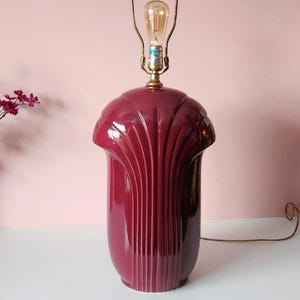 May include: A burgundy-colored ceramic table lamp with a fluted design. The lamp has a brass-colored neck and a light bulb. The lamp is on a white surface with a pink background.