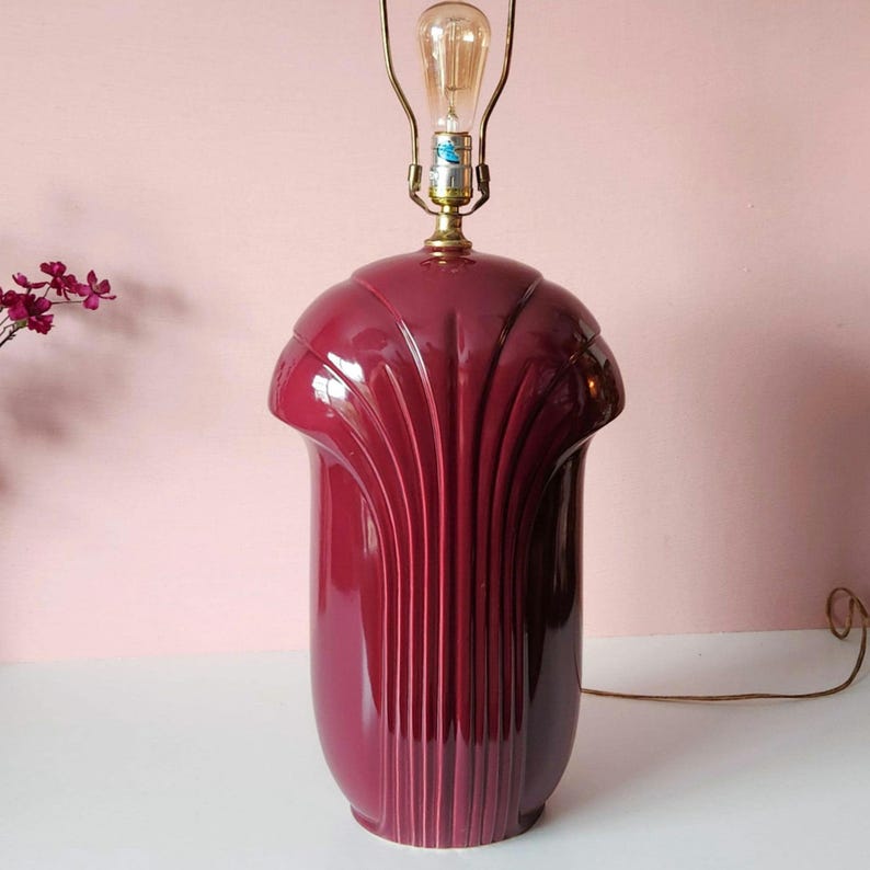 May include: A burgundy-colored ceramic table lamp with a fluted design. The lamp has a brass-colored neck and a light bulb. The lamp is on a white surface with a pink background.