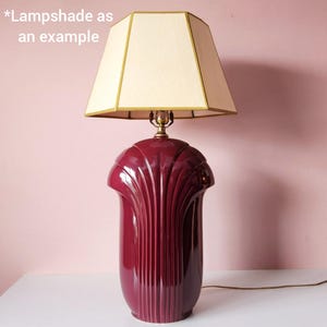 May include: A burgundy ceramic table lamp with a cream-colored lampshade. The lamp base has a fluted design and a glossy finish. The lampshade is a geometric shape with gold trim. The text "*Lampshade as an example" is in the image.