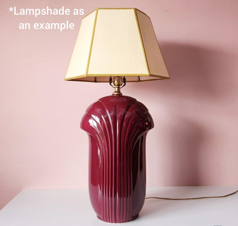 May include: A burgundy ceramic table lamp with a cream-colored lampshade. The lamp base has a fluted design and a glossy finish. The lampshade is a geometric shape with gold trim. The text "*Lampshade as an example" is in the image.