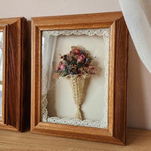 May include: A rectangular wooden frame containing a dried flower bouquet arranged in a cream-colored crocheted cone. The bouquet features pink rosebuds and assorted dried foliage, set against a plain fabric background with lace trim.