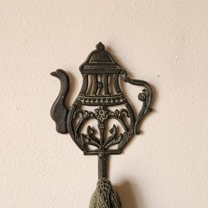 May include: A dark brown, cast iron wall hook shaped like a teapot. The metal design features an openwork pattern with a curved spout, a handle, and a central floral motif. A small portion of a textured, olive green fabric hangs from the hook.