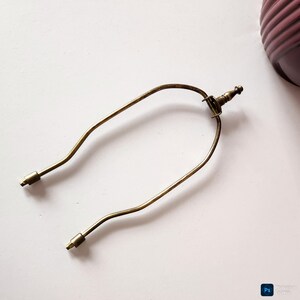 May include: A vintage-style brass lamp harp, featuring a curved design with two threaded ends for attaching to a lamp base and a finial. The harp is a warm, antique gold color.