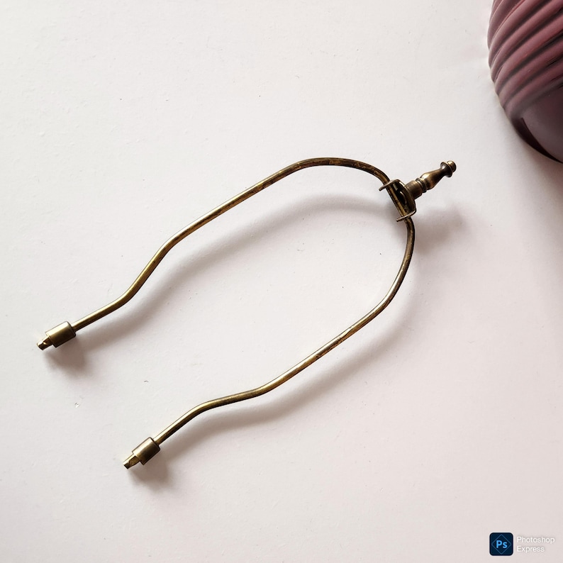 May include: A vintage-style brass lamp harp, featuring a curved design with two threaded ends for attaching to a lamp base and a finial. The harp is a warm, antique gold color.