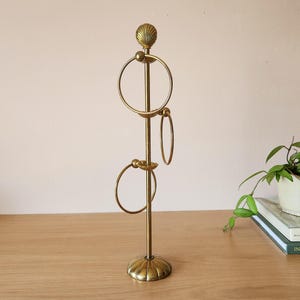 May include: A vertical brass jewelry stand featuring a central rod topped with a shell-shaped finial. The stand includes three circular rings attached at varying heights, supported by a matching shell-shaped circular base in a metallic gold finish.