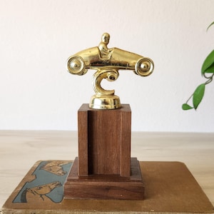 May include: A vintage trophy featuring a gold-colored race car with a driver atop a dark wooden base. The trophy has a decorative, curved support and sits on a rectangular wooden base. The trophy is a classic design.